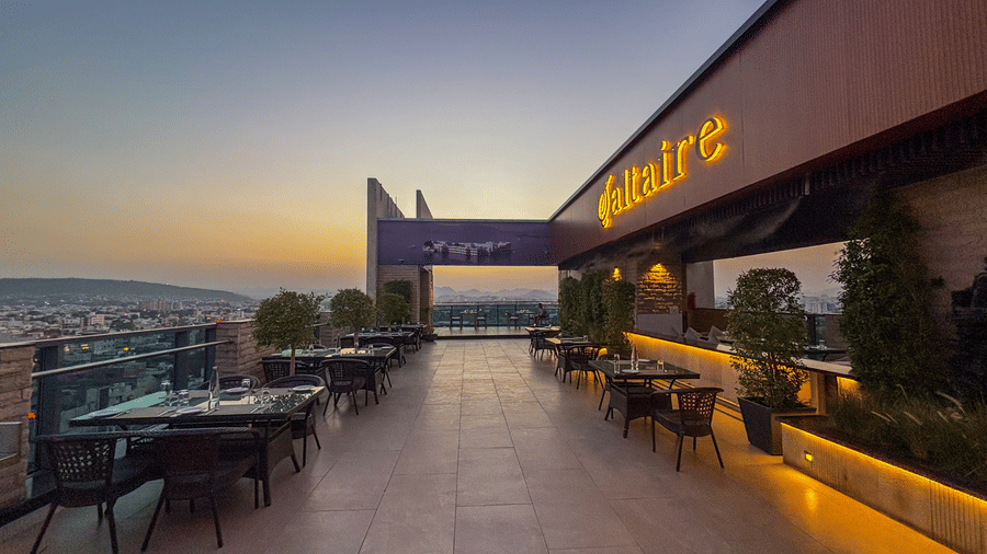 An outdoor view of the Saltaire restaurant at Pride Premier Udaipur. The terrace space features dining tables and chairs arranged along both sides of a walkway, with a structure overhead displaying signage.
