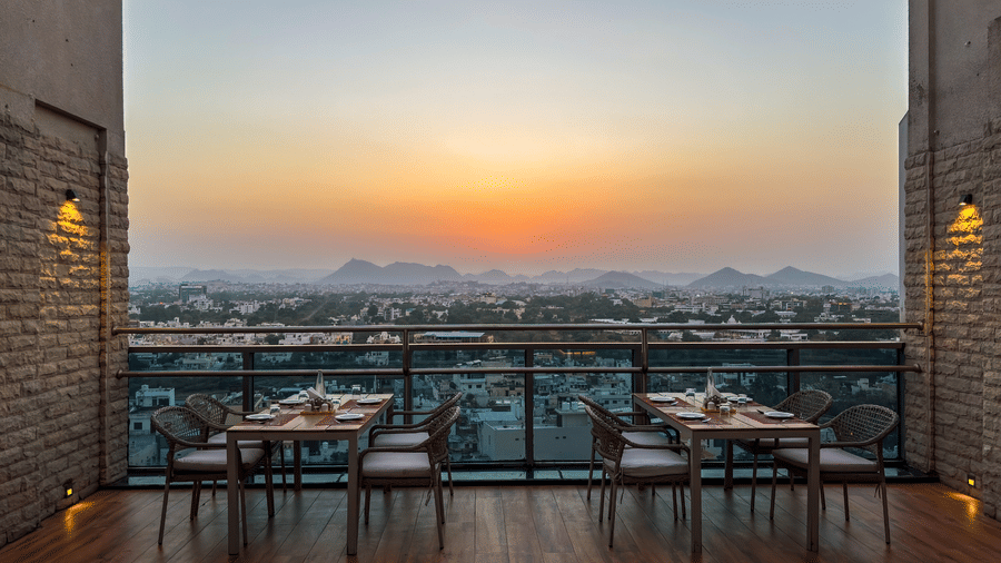 A view of a dining space at the Saltaire restaurant at Pride Premier Udaipur. Two setups of tables and chairs are positioned near a glass barrier overlooking a cityscape and distant hills.