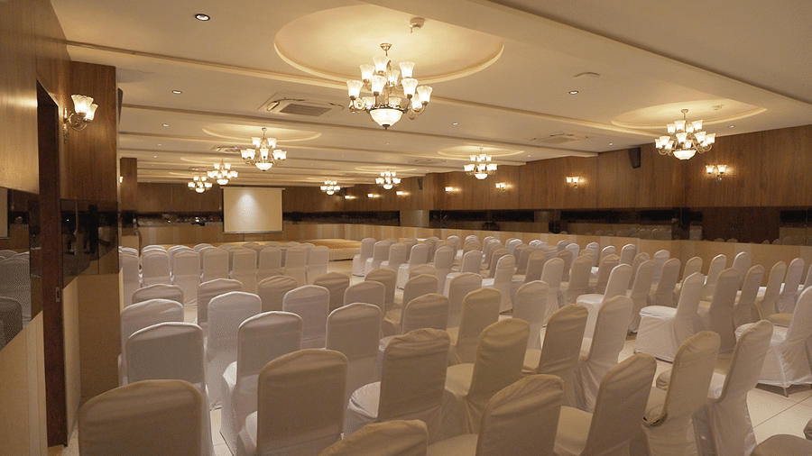 A large, elegant banquet hall set up at Pride Resort, Rajkot featuring rows of white chairs, featuring ornate ceiling lights, ready for a conference or wedding reception