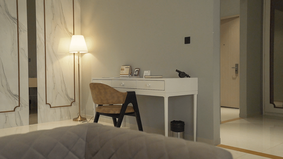 A warmly lit hotel suite room features a writing desk with a chair, lamp, and various items on top, set against a backdrop of decorative wall panels - Pride Resort, Rajkot