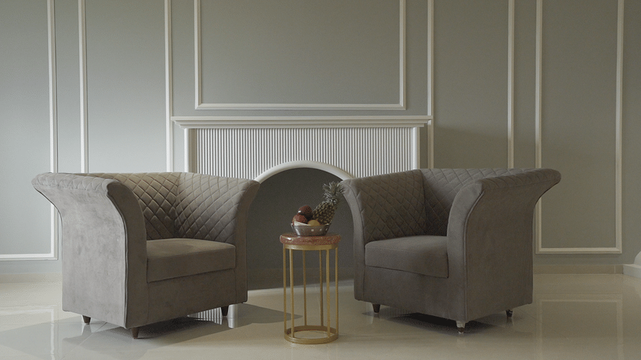 An elegant waiting area at Pride Resort Rajkot featuring two dark gray armchairs, a small gold side table, and classic wall moulding