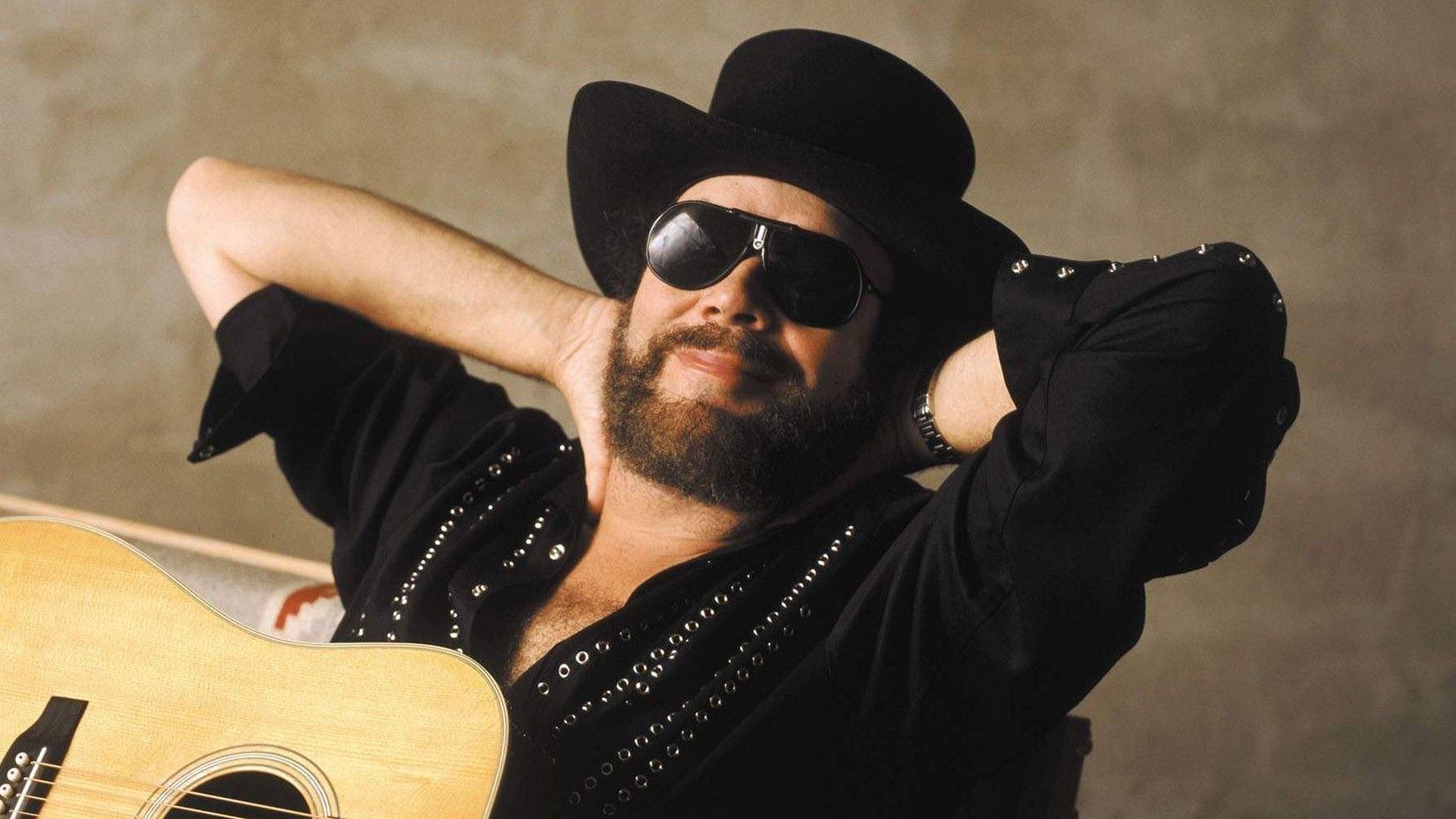 Hank Williams Jr. wearing an all black outfit, sitting relaxed in a chair with a guitar in his lap.