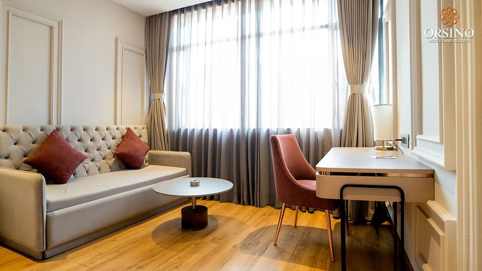 A living area at Orsino Spa Resort with a grey sofa, a small desk and chair, and large windows with curtains.