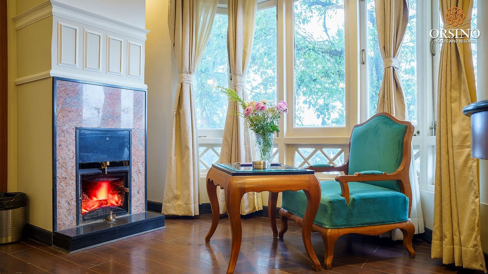 A room at Orsino Spa Resort featuring a fireplace with a lit fire, two armchairs with a small table between them, and a view through large windows.