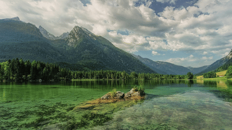 A clear green alpine lake with small rocks in the water, framed by a dense pine forest and towering, cloudy peaks.