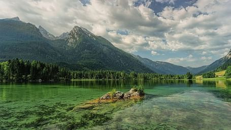 Lake reflects mountains, trees line the shore, cloudy sky above.