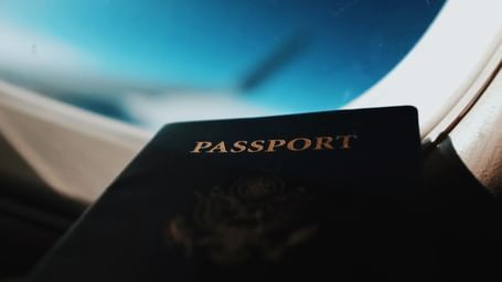 A passport rests near an airplane window, with the blurred wing and blue sky visible outside.