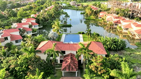 An aerial view of a large resort building with a red-tiled roof, surrounded by a swimming pool, palm trees, and a man-made lake at the Vedic Village Spa Resort.