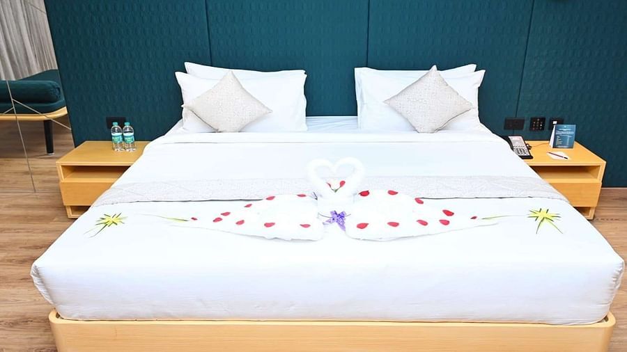 A neatly made Presedential Suite features white linens, two pillows, decorative towel swans forming a heart shape with rose petals, and bedside tables with water bottles - Dhanalakshmi Srinivasan Hotels, Perambalur
