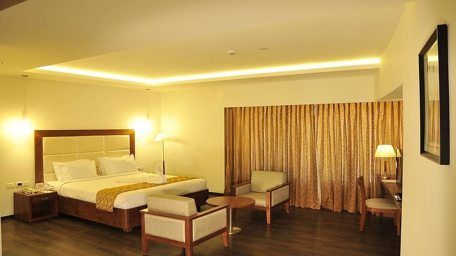 A well-lit Suite at Dhanalakshmi Srinivasan Hotels, Perambalur featuring a king size bed, two armchairs, a small table, and a work desk with a chair.
