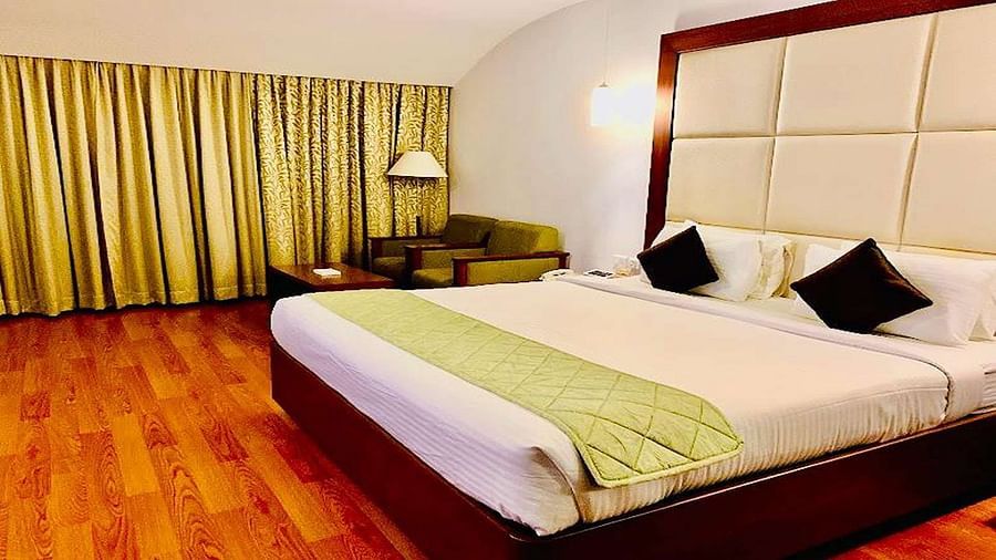 Side view of a Executive Suite at Dhanalakshmi Srinivasan Hotels, Perambalur featuring a bed with white linens, light wood flooring, and a mini-fridge.