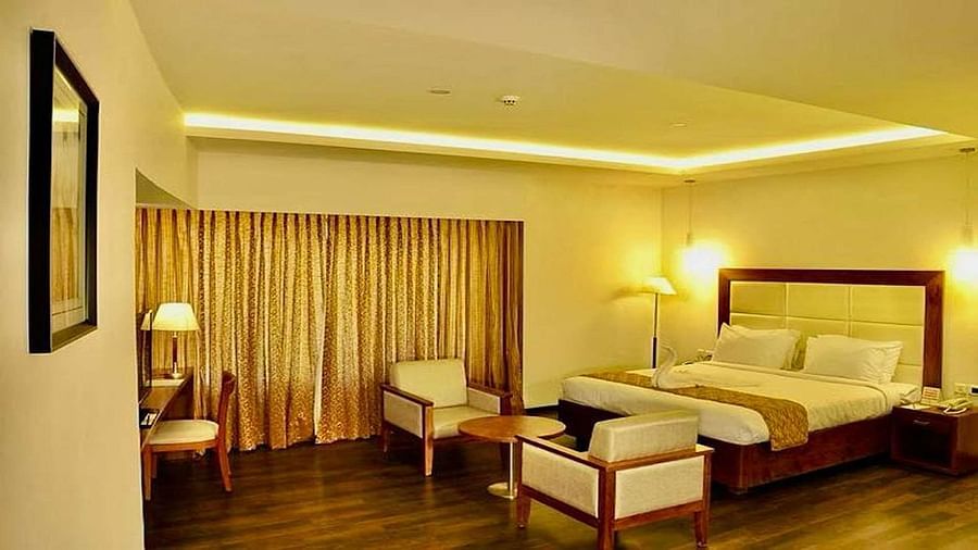 A well-lit Suite at Dhanalakshmi Srinivasan Hotels, Perambalur featuring a king size bed, two armchairs, a small table, and a work desk with a chair.