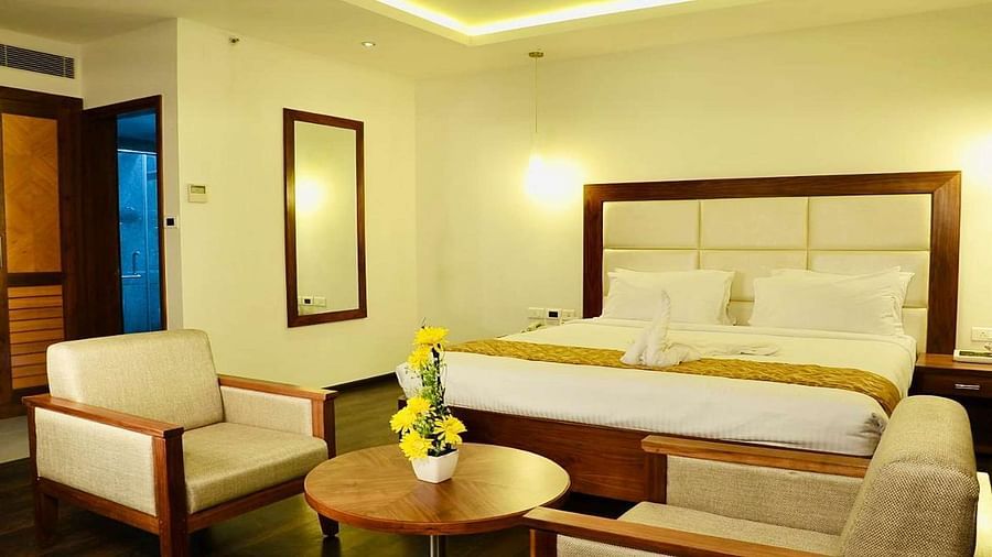 A close up view of well-lit Suite at Dhanalakshmi Srinivasan Hotels, Perambalur featuring a king size bed, two armchairs, a small table, and a work desk with a chair.