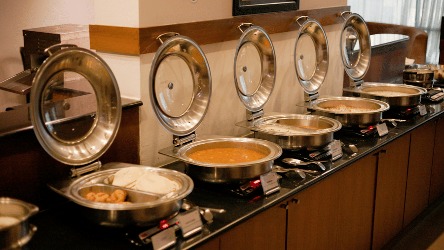 A row of silver chafing dishes with open lids containing various foods on a dark counter in a dining area at Hotel Horizon, Trivandrum.