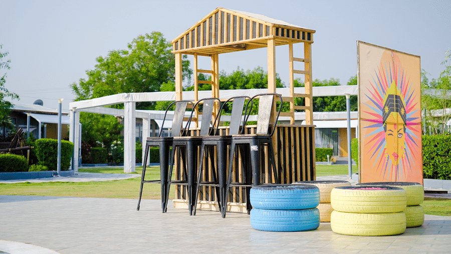 A shaded outdoor seating space with high tables and stools beside a landscaped lawn.