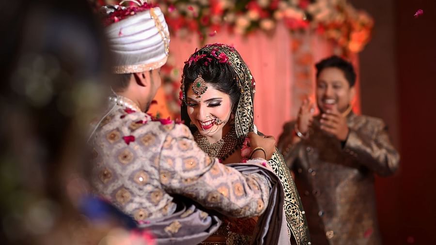bride and groom standing together with a person clapping behind them - Rhythm Gurugram