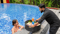 Resort staff serving a floating tray of food and cocktails to a guest relaxing at the edge of a blue swimming pool.