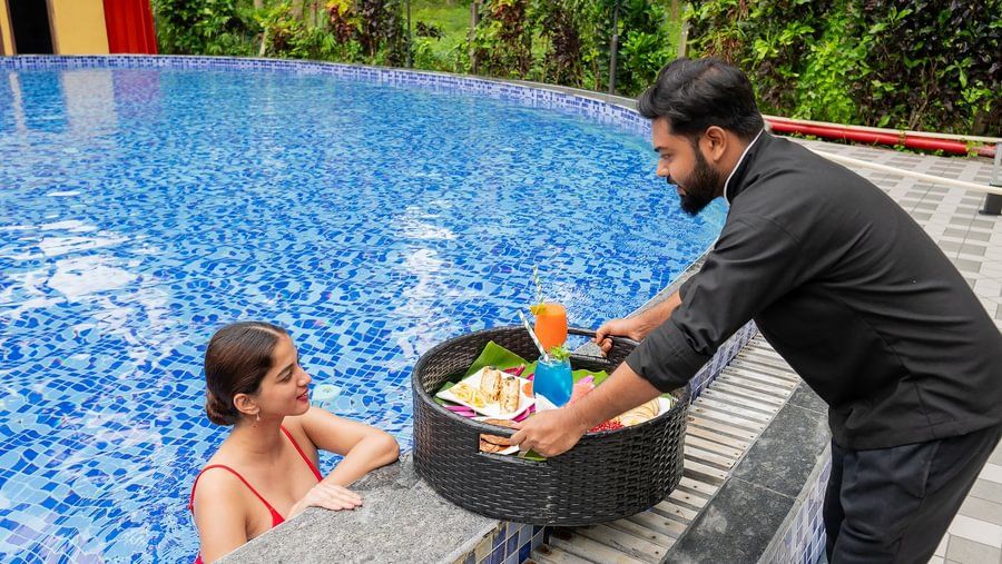 Resort staff serving a floating tray of food and cocktails to a guest relaxing at the edge of a blue swimming pool.