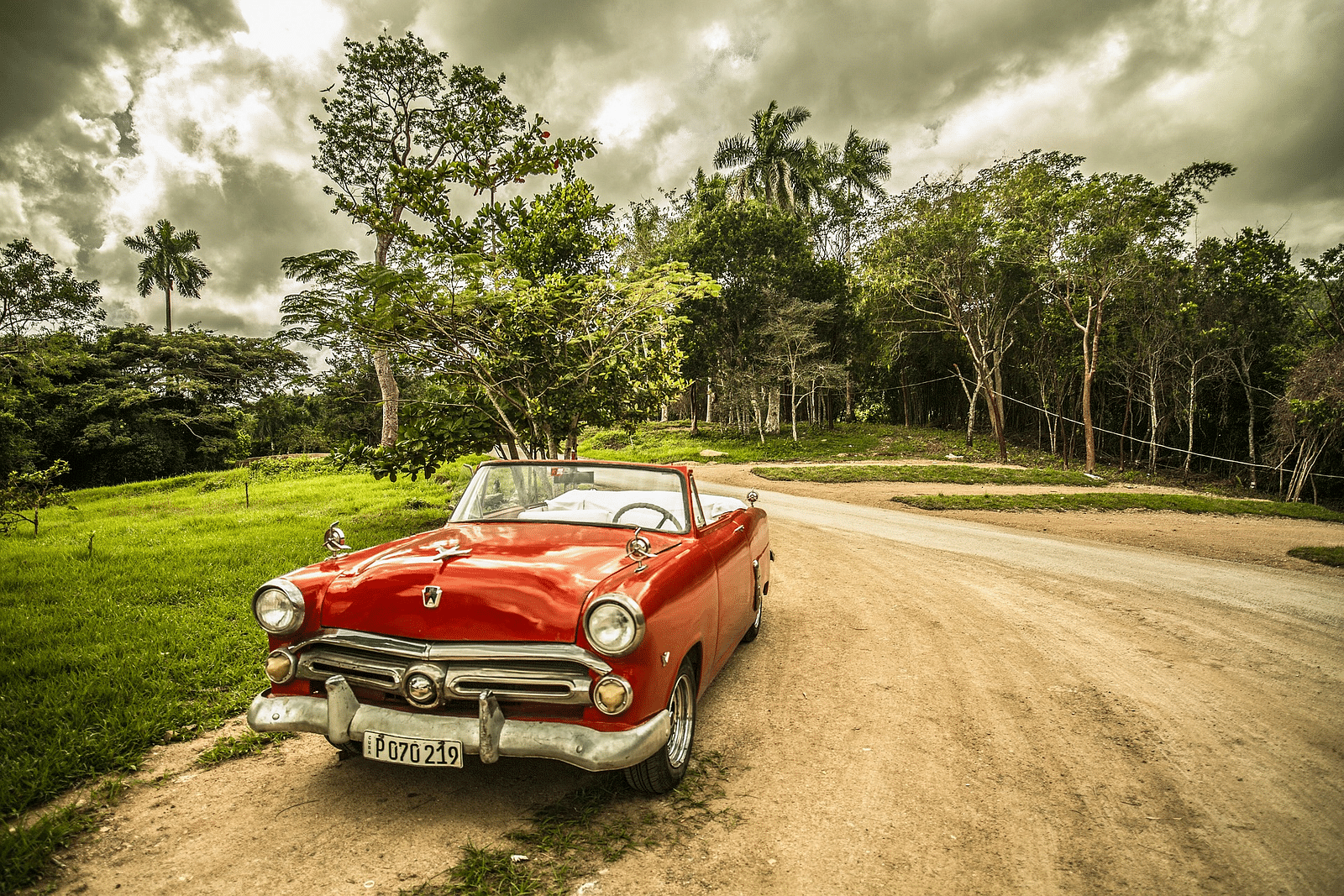 An vintage luxury open top car in red colour parked on a mud road with trees in the background.