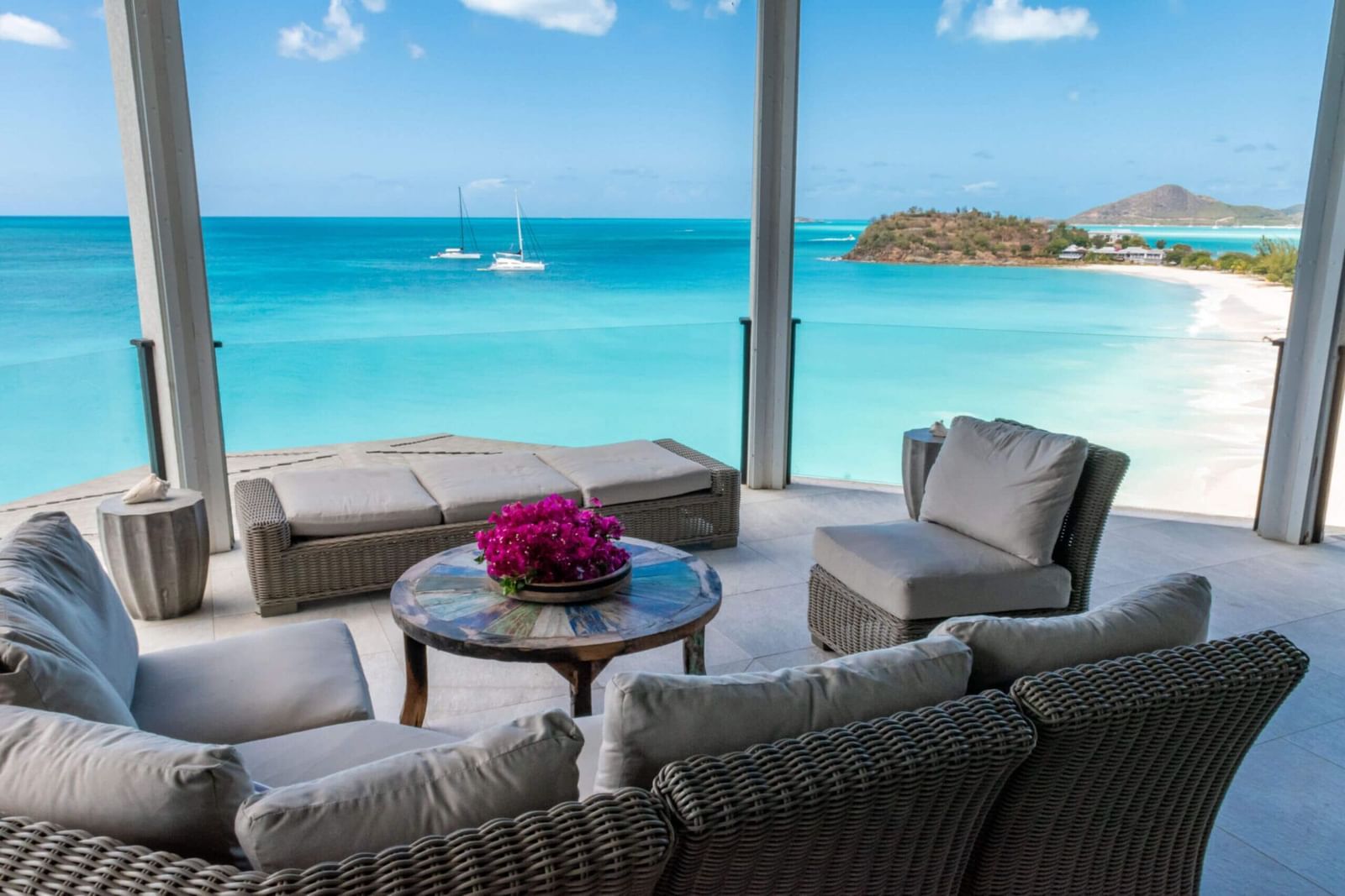  Balcony lounge area overlooking a pristine tropical beach with calm waters and distant sailboats.
