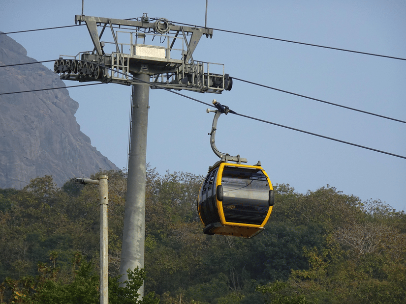 A yellow and black cable car suspended from a metal pylon, moving across a wooded hillside under a pale sky.