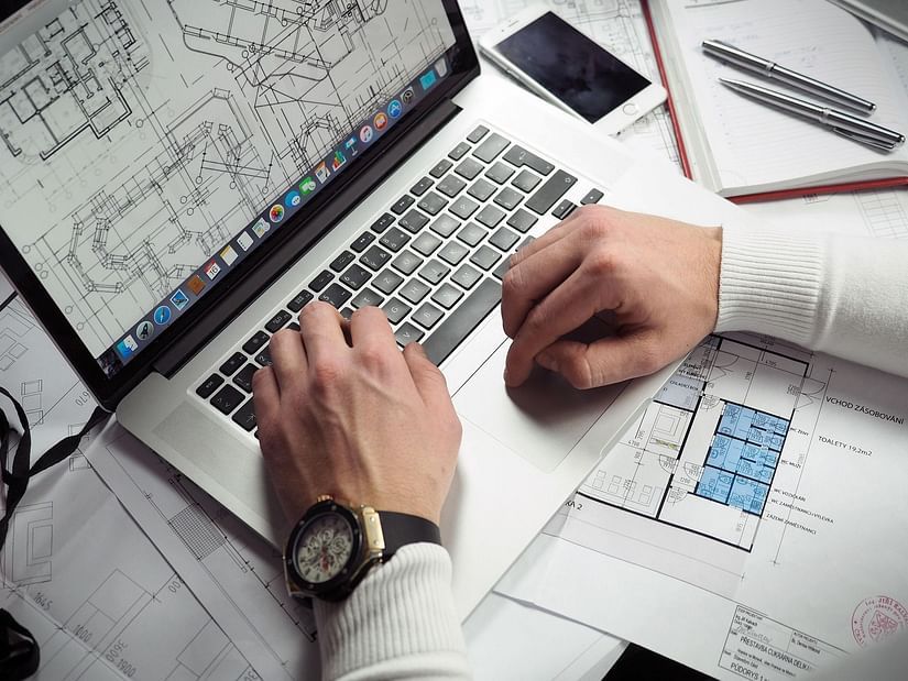 Hands of a man in formal attire working on a laptop, surrounded by papers.