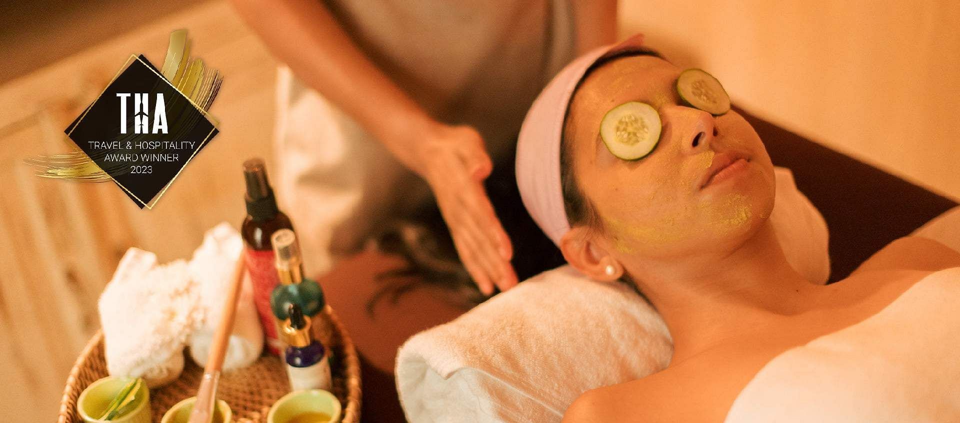Person receiving a facial treatment with cucumber slices on eyes; massage oils and towels displayed at Karma Chalets.