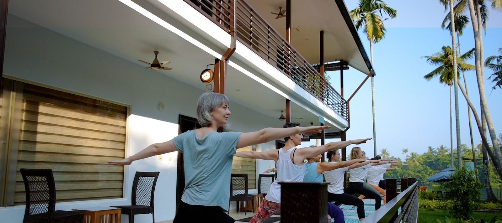 A group of people practice yoga warrior pose on a patio with a building and palm trees in the background.