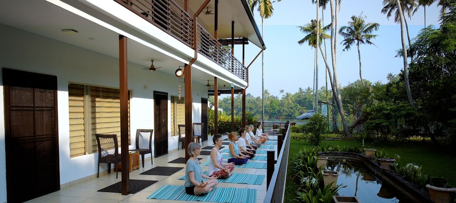 A group of people sit in a row on yoga mats, practicing meditation or yoga on a long patio with a building and tropical foliage in the background.