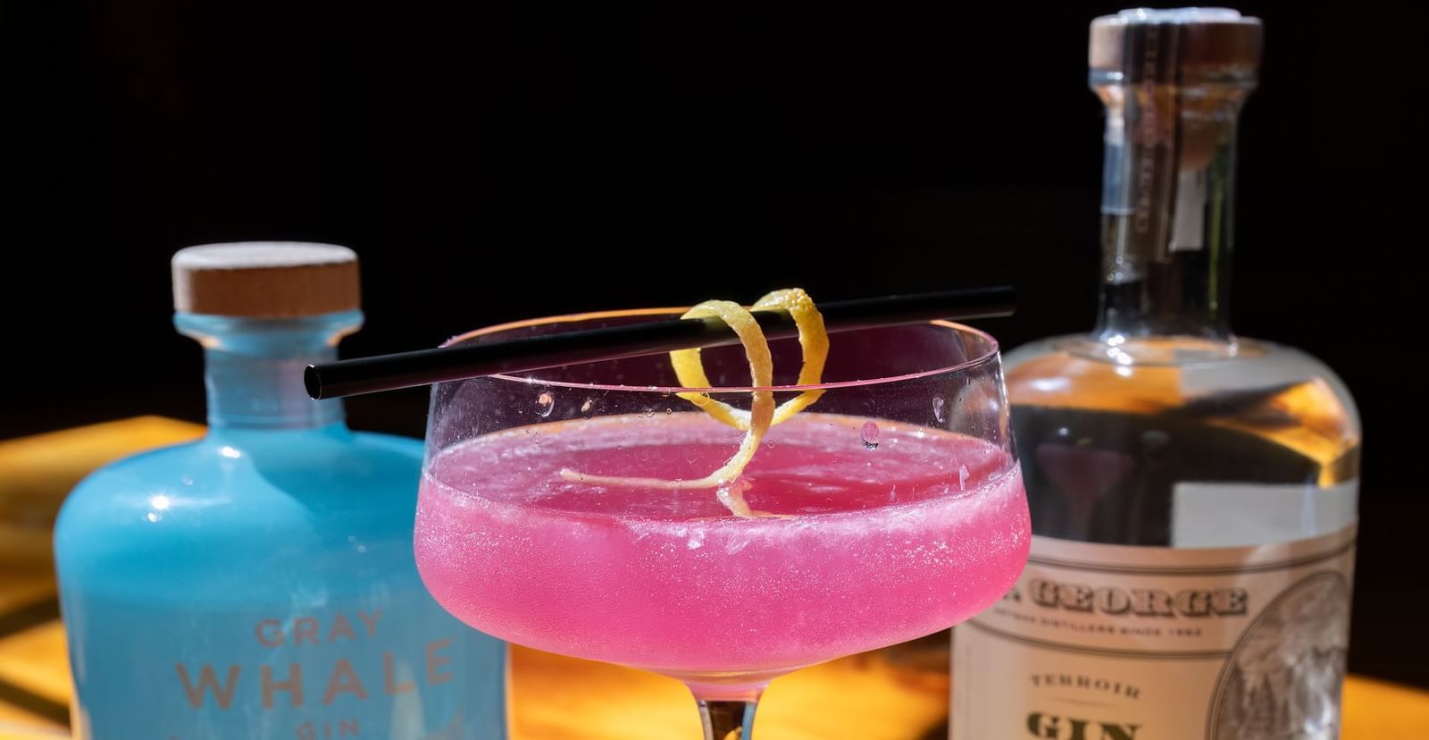 Two vibrant cocktails with lemons and gin bottles on a wooden bar under warm lighting at Narrow Gauge Inn.