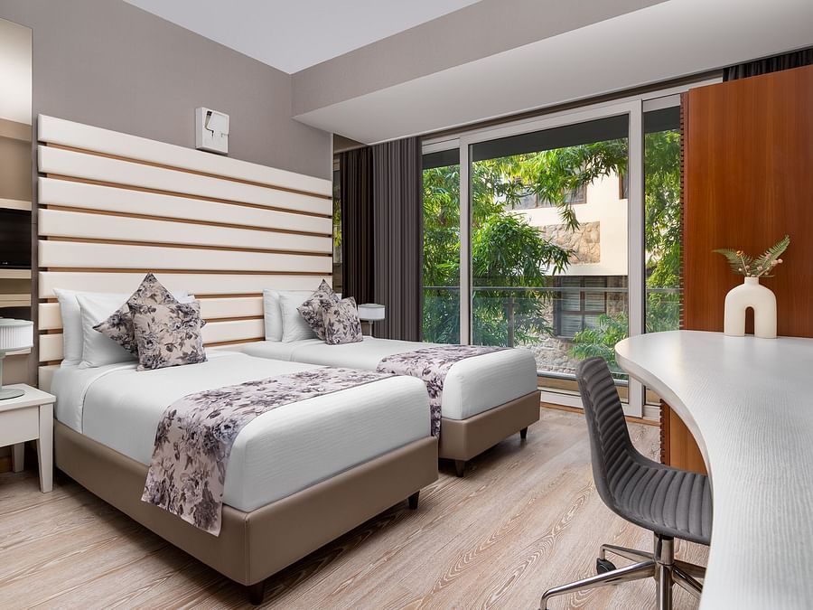  Contemporary room at The Atara, Gurugram featuring two single beds, a white desk, and a balcony view. The room has light wood floors and horizontal wall decor