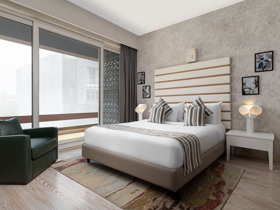 A stylish room at The Atara, Gurugram boasts a comfortable bed with a patterned runner, a plush green armchair, and a large window that offers a scenic outdoor view