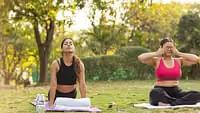 Two women in two different yoga postures on yoga mats in the lawn area surrounded by trees and plants at Daksh Eden Greenz, Sasan Gir