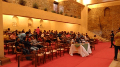 A large group of people are seated in chairs on a red carpet, facing a table covered with a white and green cloth, inside what appears to be a hall or event space at Tijara Fort-Palace - 19th Century, Alwar.