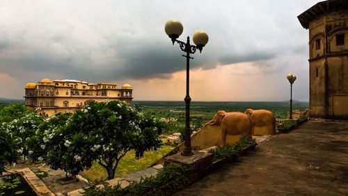 A view of Tijara Fort-Palace in Alwar shows its architecture, surrounding greenery, and an overcast sky.