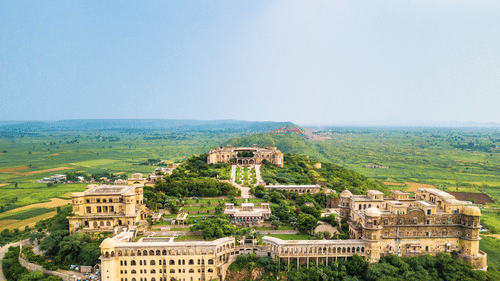 An aerial view of Tijara Fort Palace, perched on a hilltop with greenery surrounding it.