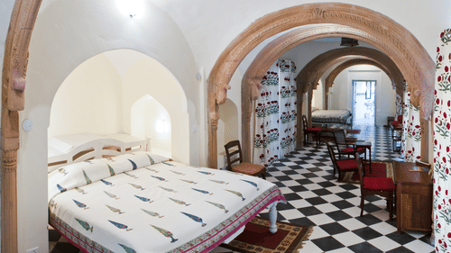 A suite at Tijara Fort-Palace - 19th Century, Alwar showcases a bedroom with a bed featuring white linens with a colorful pattern, a bedside table, and arched doorways leading to a dining area with a table and red chairs.