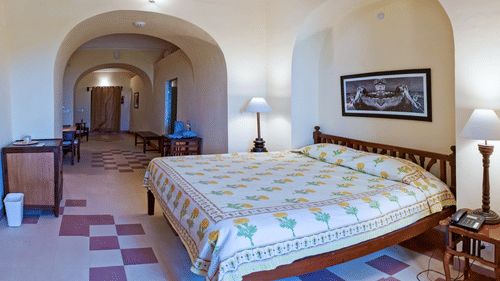 A suite at Tijara Fort-Palace - 19th Century, Alwar features a bedroom with a bed with floral linens, bedside tables with lamps and a telephone, and framed artwork, connected by arched doorways to a living area with a desk and seating.
