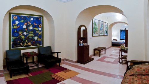 A common area at Tijara Fort-Palace - 19th Century, Alwar features arched openings, tiled flooring with colorful rugs, seating areas with black chairs and a small table, a wooden stand with amenities, framed artwork on the wall, and a glimpse into a bedroo