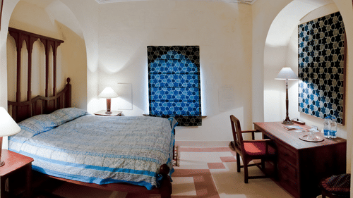A guest room at Tijara Fort-Palace in Alwar features a bed with blue and white striped linens, bedside tables with lamps, a desk with a chair and a lamp, decorative blue textiles on the wall, and tiled flooring.