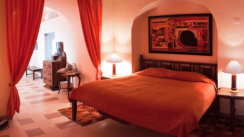A guest room at Tijara Fort-Palace - 19th Century, Alwar features a bed with orange linens, bedside tables with lamps, artwork above the bed, and an arched opening with red curtains leading to another part of the room with a dresser and seating.