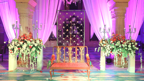 A decorated seating area with a wooden chair, floral arrangements, and purple-lit ornate backdrop is prepared for an event at Tijara Fort-Palace - 19th Century, Alwar.