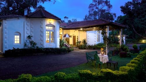 Facade of the hotel in the night with metal chairs and a small table in the garden - Wallwood Garden - 19th Century, Coonoor