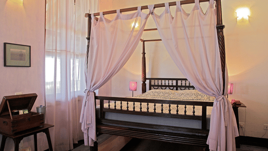 An elegant 4-poster bed with light coloured drapes, a double mattress, and classic wooden furniture in the bright, heritage-style Prince Christian - Sea View room at The Bungalow on the Beach, Tranquebar.