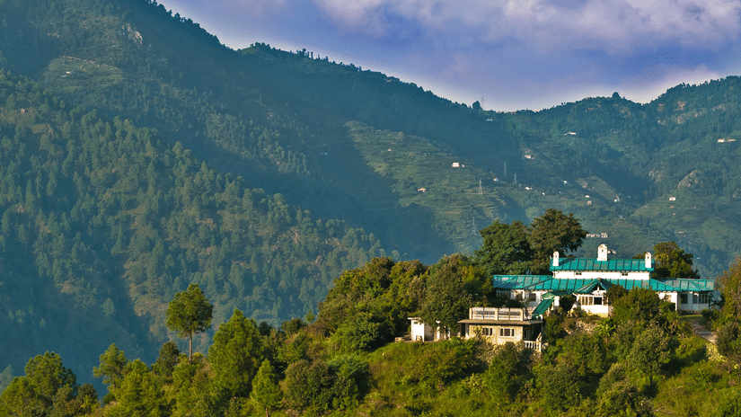 Facade of The Ramgarh Bungalows perched on a hill surrounded by scenic mountain view captured during the day.