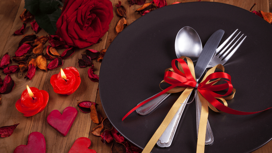 Romantic Valentine table setup with black plate, cutlery tied with red ribbon, roses and heart decor.