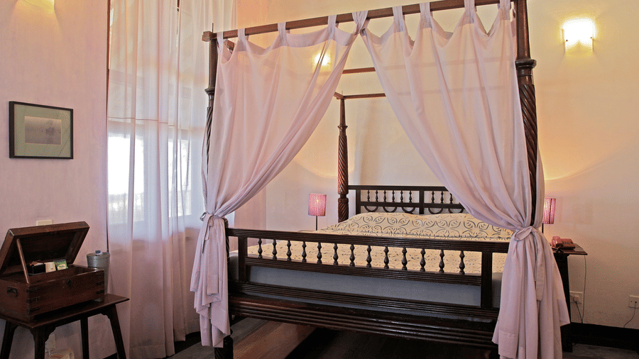 An elegant 4-poster bed with light coloured drapes, a double mattress, and classic wooden furniture in the bright, heritage-style Prince Christian - Sea View room at The Bungalow on the Beach, Tranquebar.