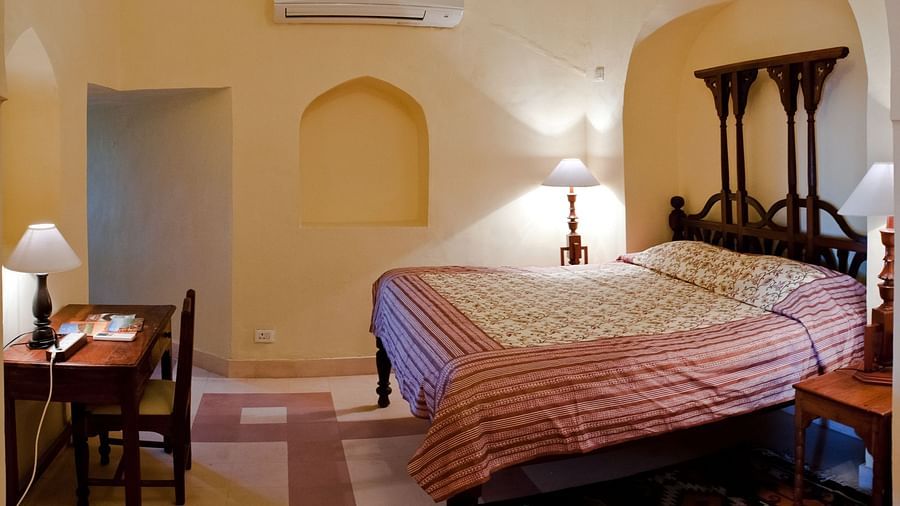 A guest room at Tijara Fort-Palace - 19th Century, Alwar features a bed with patterned linens and a striped throw, bedside tables with lamps, a desk with a lamp and chair, an arched niche in the wall, and air conditioning.