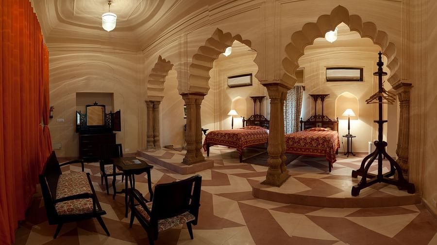 A spacious room showcases two beds with bedside tables and lamps, separated by ornate arched pillars, along with seating chairs and other furniture at Tijara Fort-Palace - 19th Century, Alwar.