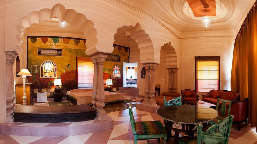 A spacious interior shows multiple seating areas with chairs and sofas, along with beds and arched architectural elements inside Tijara Fort-Palace - 19th Century, Alwar.